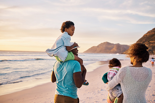 Rear view of a carefree african american family walking and having fun together on the beach. Parents spend time with their sons during sunset. Little siblings playing with their parent's vacation