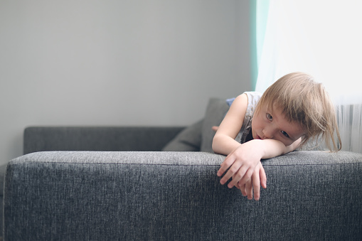 European toddler child sits and gets bored on gray sofa in bright living room, minimalist interior