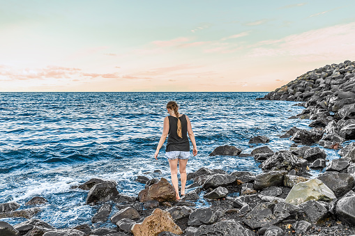 Young adult girl enters the Atlantic Ocean on the Playa Negro beach in Santa Cruz de Tenerife, Spain - view from the back. Barefoot woman in shorts and a T-shirt stands among large stones in sea water