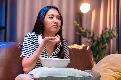 ad young asian woman watching television and crying while still eating popcorn on sofa relax before sleep in late night. upset lady holding snack watch tv in dark bedroom in midnight lifestyle