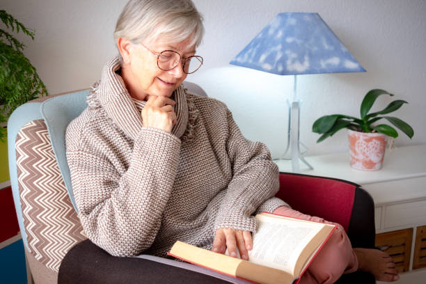 自宅で肘掛け椅子に座って本を読んでいる美しいリラックスし��たシニア女性の肖像画 - 古い世代と退職のコンセプト - old armchair women senior adult ストックフォトと画像