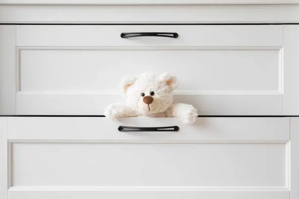 Photo of Smiling white teddy bear looking from opened drawer at home. Closeup. Front view.
