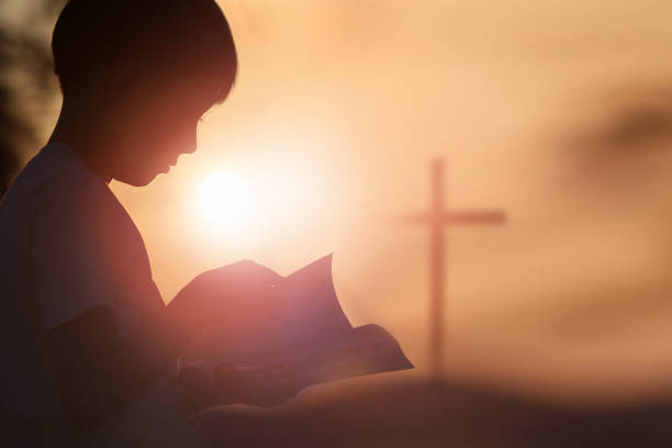 a child reading a bible and a cross in the sunset sky - color image jesus christ child people imagens e fotografias de stock
