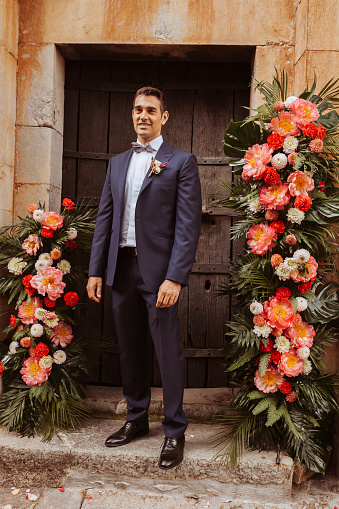 Young happy handsome groom posing with suit in front of the church with floral bouquets.\nWedding day, human relationship, engagement, newlyweds.