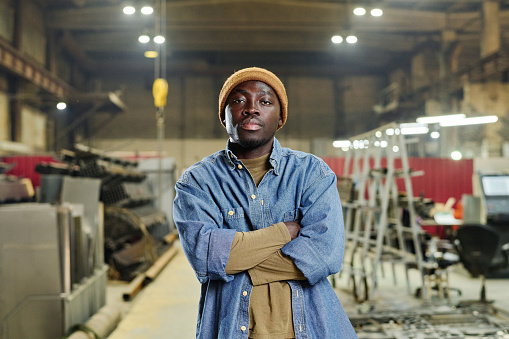 Portrait of African young worker in casual clothing standing at factory with his arms crossed and looking at camera
