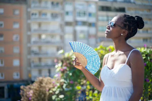 Young Woman wearing sunglasses  fanning herself  using a blue hand fan on a hot day  with a Blurred city view background.