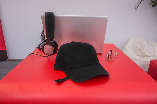 Pink laptop, black headphones, golden glasses and a black cap on a red table with copy space. On the right there are two soft seats. The cap can have your company logo on it.