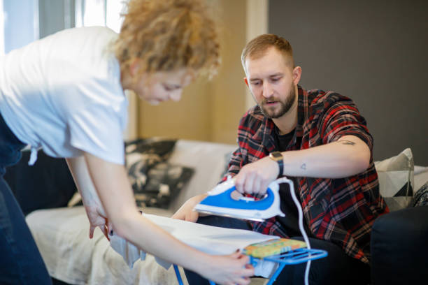 Young man sitting on the sofa in the living room and ironing on the board, his flatmate helping him Young man sitting on the sofa in the living room of a shared apartment and ironing his clothes on the board, his flatmate standing by and tightening shirt to help, sharing chores in daily routine flatmate stock pictures, royalty-free photos & images