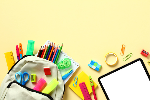 Backpack with colorful school supplies on yellow table, top view. Back to school concept.