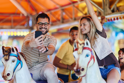 Young happy friends having fun while taking a selfie with cell phone during carousel ride at amusement park.