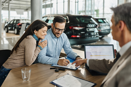 Happy couple signing a contract for buying a car on a meeting with salesman in a showroom.