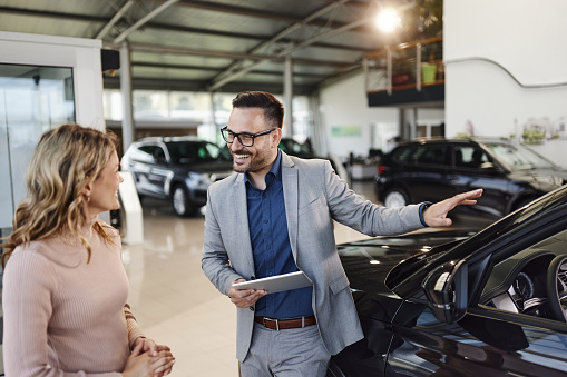Happy car salesman talking to his customer while showing her a car in a showroom.
