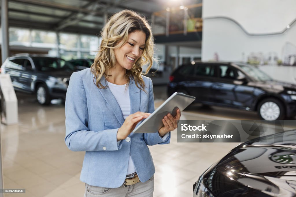Happy car salesperson using digital tablet in a showroom. Happy female salesperson working on touchpad in a car showroom. Car Dealership Stock Photo