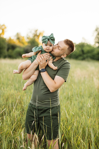 Portrait of young man smiling and looking at camera while walking in the meadow during picnic with his daughter on hands. Father enjoying every moment spending with his loving child.