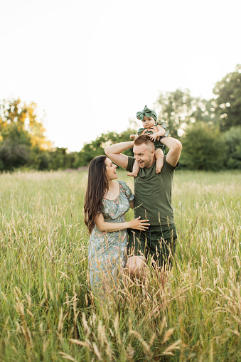 Beautiful caucasian family of father, mother and little daughter posing on camera on background of green field. Dad holds daughter on his shoulders. Concept of parenthood, happiness and leisure time.