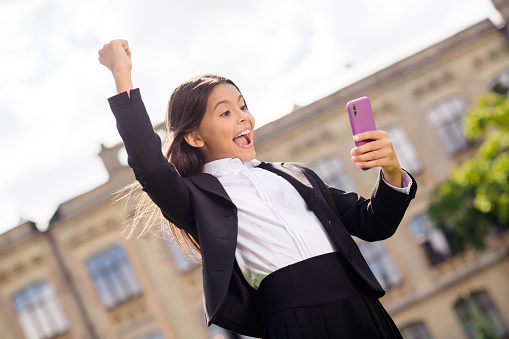 Photo of little pupil girl hold cellphone raise fist cheerful look screen wear black uniform white shirt urban city outdoors.