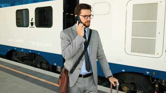 Mid adult businessman with luggage talking on his smart phone at a railroad station