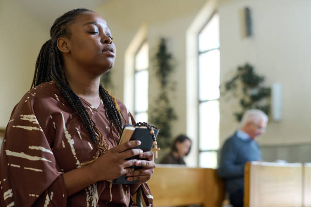 Young black woman in casual dress keeping her eyes closed during prayer Young black woman in casual dress keeping her eyes closed during silent prayer in church while holding Holy Bible and rosary beads church ladies stock pictures, royalty-free photos & images