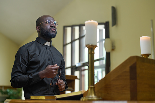 African American priest in black casualwear holding rosary beads during pray while standing by pulpit with two burning candles