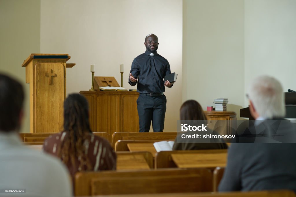 Confident priest of evangelical church with Bible in hand saying sermon Confident priest of evangelical church with Holy Bible in hand saying sermon while standing in front of intercultural parishioners Pastor Stock Photo