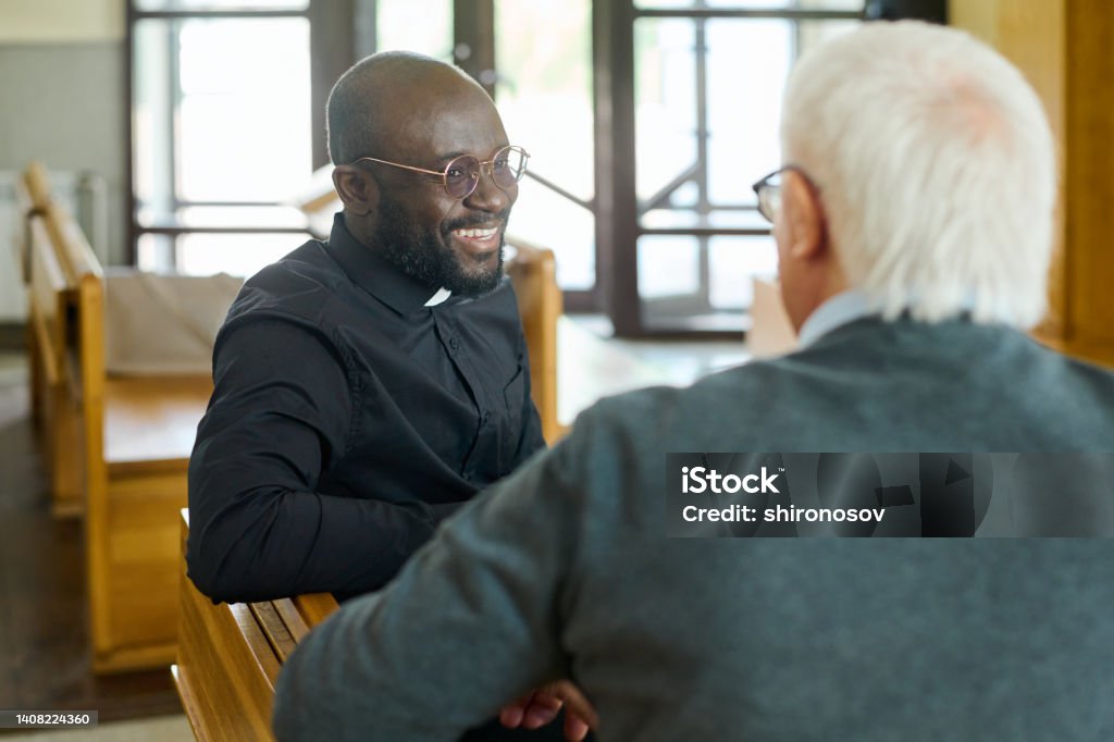 Happy young African American pastor of evangelical church consulting aged man Young cheerful African American pastor of evangelical church consulting aged grey haired male parishioner after sermon Priest Stock Photo