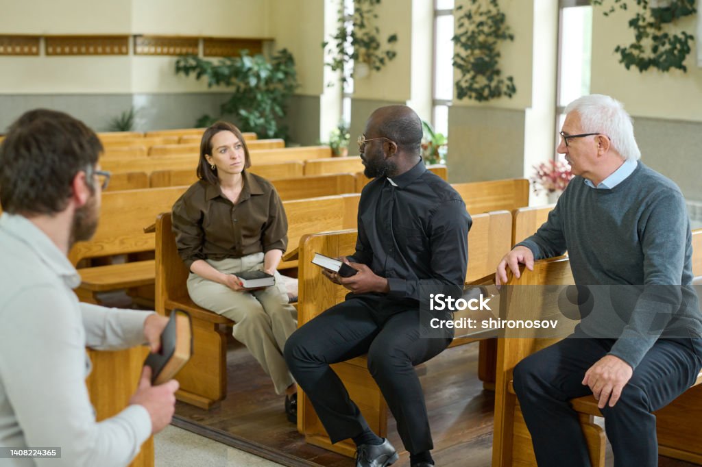 Young African American priest with Bible talking to one of parishioners Young African American priest with Bible talking to one of parishioners while explaining her some verses from New Testament Church Stock Photo