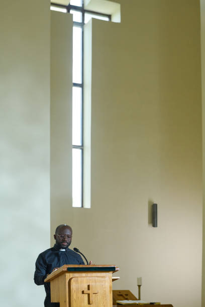 istock Young black man standing by wooden pulpit while reading Bible aloud 1408224368