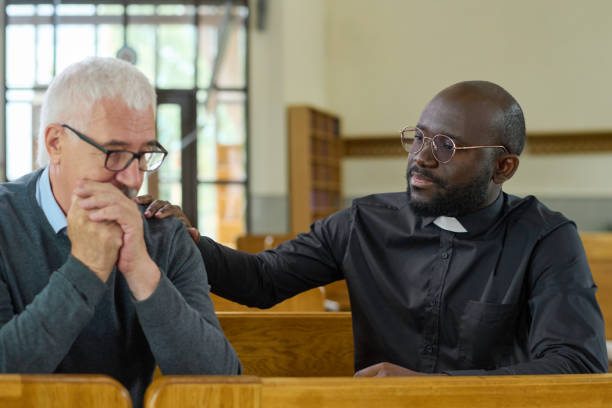 Black man in shirt with clerical collar keeping hand on shoulder of parishioner Young black man in shirt with clerical collar keeping hand on shoulder of senior male parishioner while comforting him and praying clergy stock pictures, royalty-free photos & images