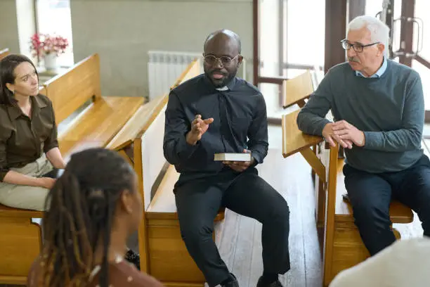 Young confident pastor of evangelical church explaining something to woman sitting in front of him during gathering after sermon