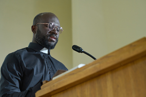 Joven sacerdote de camisa negra con cuello clerical pronunciando discurso por púlpito photo