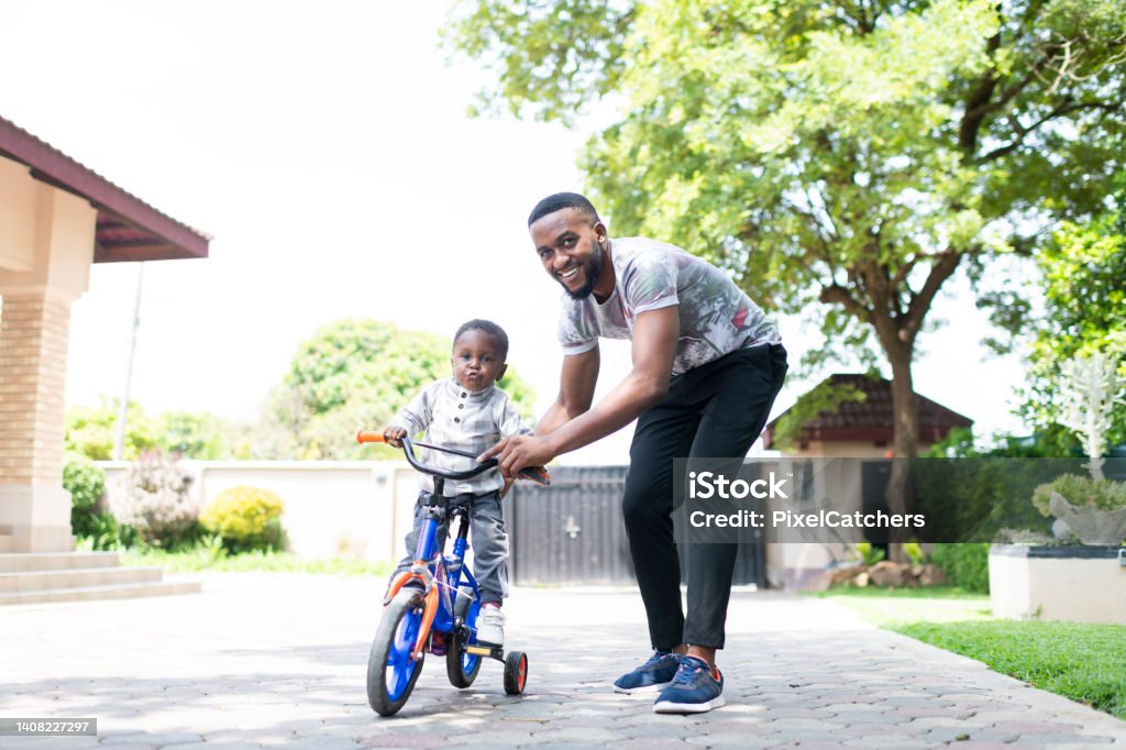 Young father helps toddler ride tricycle in driveway of home Young father helps toddler ride tricycle Front Yard Stock Photo