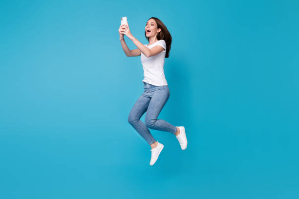 Full length body size view of her she attractive pretty slim fit funky cheerful girl blogger jumping having fun using cell app 5g fast speed isolated bright vivid shine vibrant blue color background stock photo