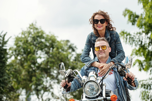 Cool happy mature couple in demin jackets riding motorbike in city