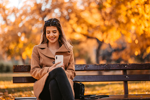 Beautiful young woman sitting on a bench in the public park during autumn and using smart phone.
