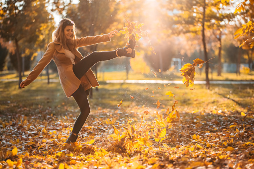 Beautiful young woman kicking a heap of leaves in the park as the sun sets.
