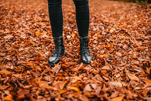 Low section of a young woman standing on dry autumn leaves in the park during autumn.