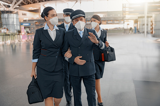 Male pilots and female flight attendants wearing pronectice face masks and walking in airport terminal together