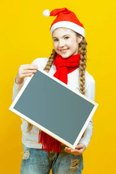 Happy Winsome Caucasian Teenager Girl In Santa Hat and Red Scarf  Holding Black Wooden Blackboard While Pointing With Pointfinger On Yellow Background. Vertical Composition