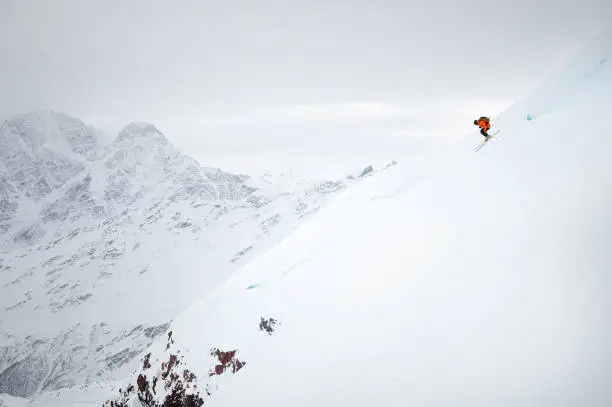 A male skier quickly skis down a snow-covered glacier, against the backdrop of high mountains and a cloudy sky