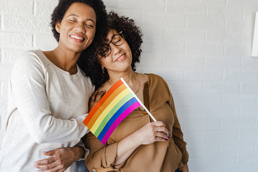 Portrait of African American woman leaning on her girlfriend against white wall and holding LGBTQIA flag. They are having eyes closed.