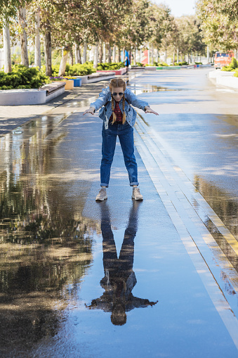 A woman in a denim outfit stands with her body tilted and her arms outstretched forward in a wet alley in a park. Walk after the rain. Cheerful image.