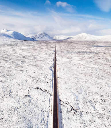 An aerial view of a long, straight and empty road leading towards the snow-covered mountains of Glencoe on the horizon. Photographed in February.
