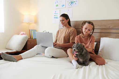 Young woman working on laptop in bed when daughter playing little dog bear by