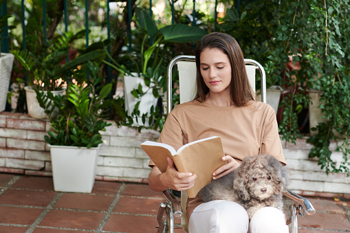 Woman sitting on chair in backyard with dog on her laps and reading book