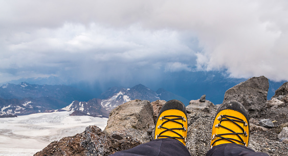 First person view to legs in yellow plastic climbing boots takes a rest on the top of the mountain