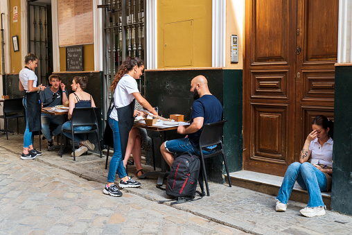 Seville, Spain -- May 15, 2022. Photo of patrons dining at an outdoor restaurant in Seville; a young lady sits in a doorway.