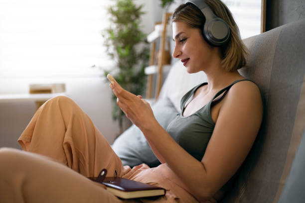 Caucasian woman sitting on the sofa, using mobile phone while listening to music on wireless headphones stock photo