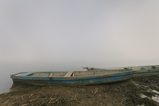 Two old blue weathered shabby retro boats on the riverbank in thick fog. Cottagecore or unplugged concept. Countryside calm soothing landscape. Copy space. Horizontal orientation.