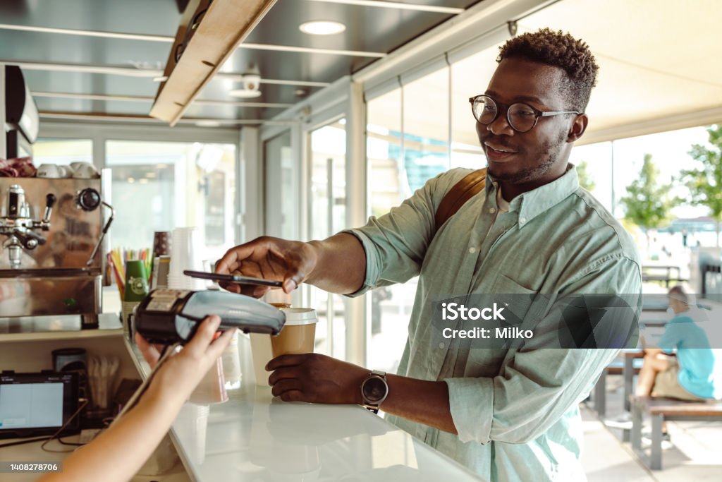 An African American young man paying at the coffee shop Young African American student paying a coffee with his phone Paying Stock Photo