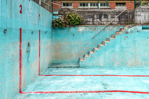Abandoned swimming pool, focus on corner with stairs and changing rooms in the background. The pool has a bright blue colour with red stripes.